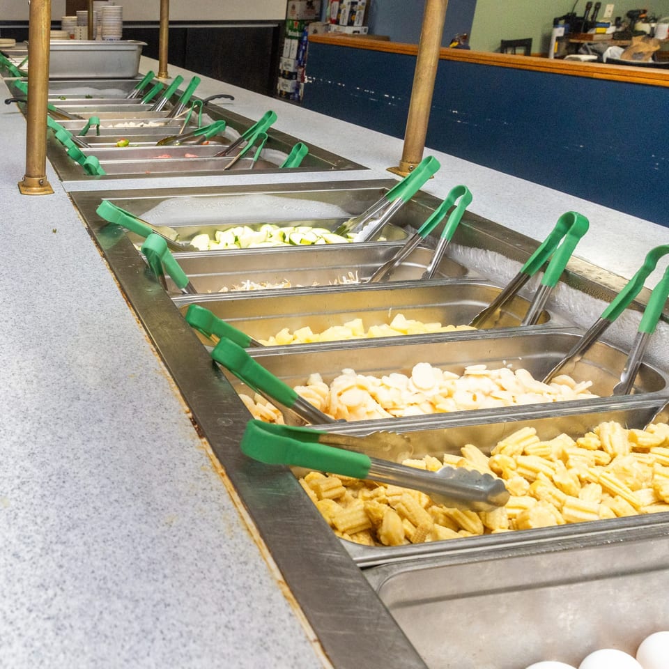 A buffet line at Hi Ho Mongolian Grill with various chopped vegetables and tongs in metal bins.