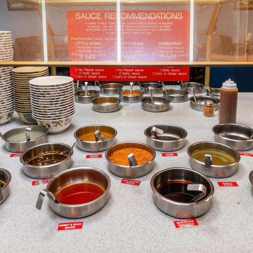 A buffet-style sauce bar at Hi Ho Mongolian Grill, with various sauces in metal bowls and a sauce recommendation sign.
