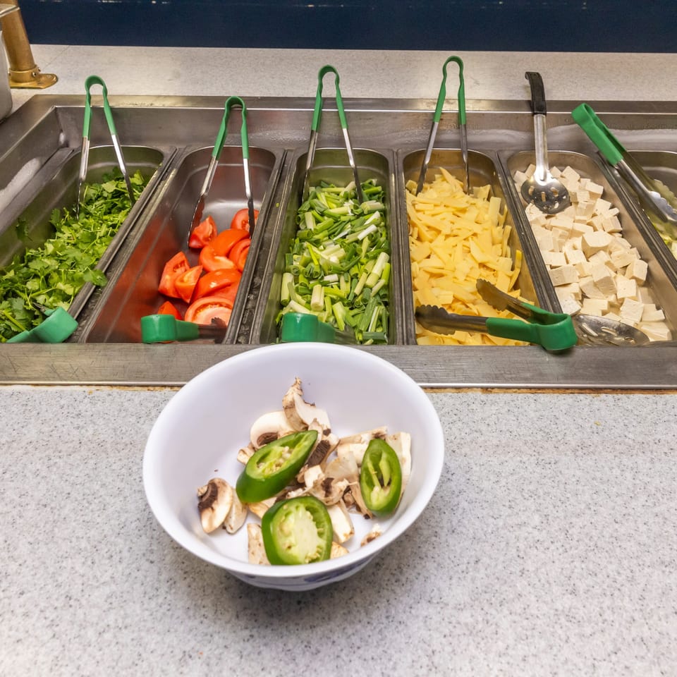 A Mongolian grill buffet line with fresh cilantro, tomatoes, green onions, bamboo shoots, and tofu.